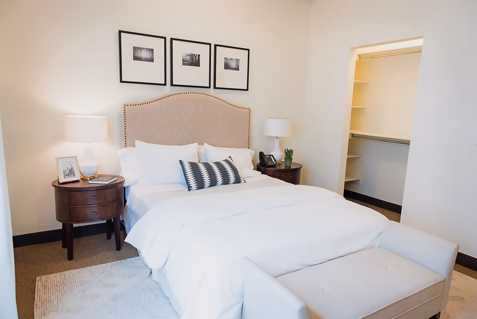 A neatly made bed with white bedding and a beige upholstered headboard in a bedroom. There are two dark wooden nightstands on either side of the bed, each with a white lamp. One nightstand has a framed photo and a book, while the other has a small plant and a phone. Above the bed are three black and white framed pictures. To the right, there is an open walk-in closet with shelves and hanging rods. A light-colored bench is placed at the foot of the bed.