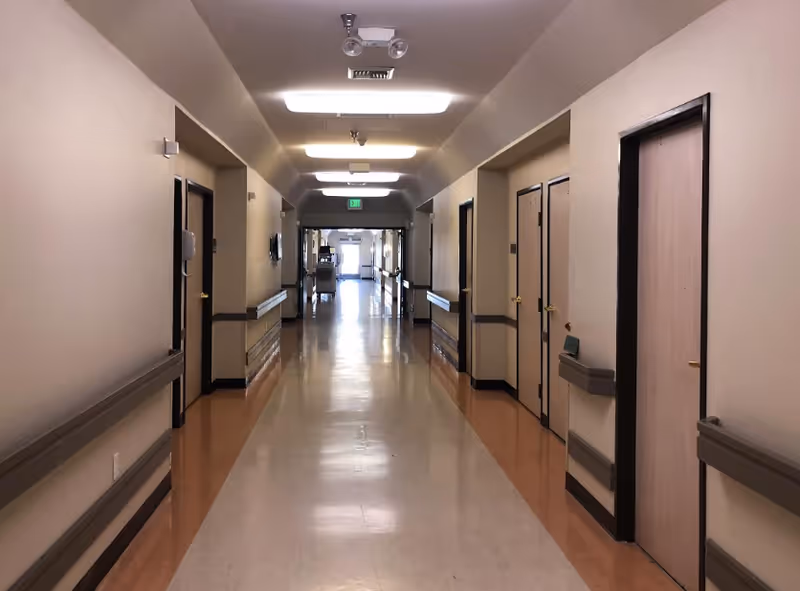 Long, empty nursing home hallway with doors on both sides, handrails, and overhead lights.