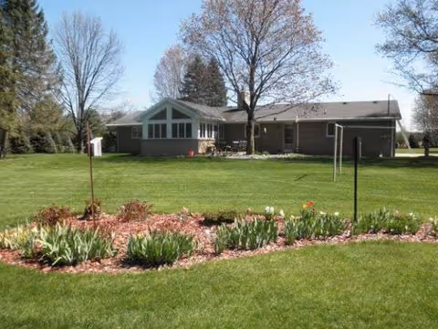 Single-story senior living facility with a manicured lawn, flower beds, and leafless trees.