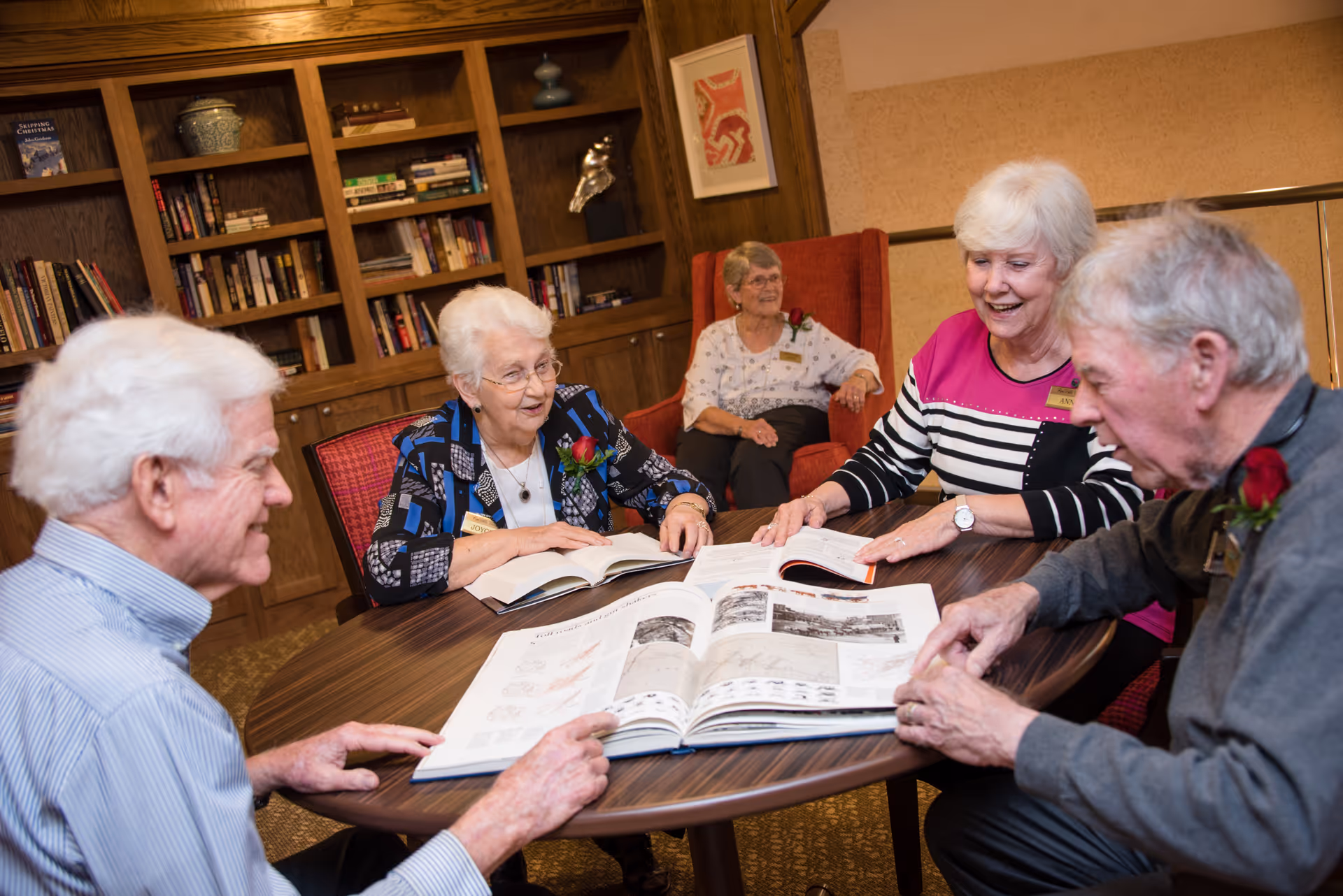 Four elderly individuals sitting around a round wooden table looking at open books, with a fifth elderly person seated in a red armchair in the background. Behind them is a wooden bookshelf filled with books and decorative items.