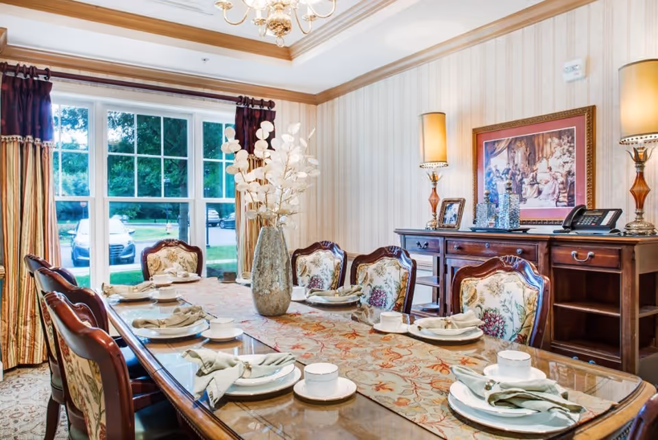 A formal dining room with a long wooden table set for six people. The table has a floral table runner, white plates, cups, and neatly folded napkins. The chairs have floral upholstery. A large vase with white decorative branches is placed in the center of the table. Behind the table is a wooden sideboard with two lamps, framed pictures, and a phone. Large windows with curtains allow natural light to enter the room.