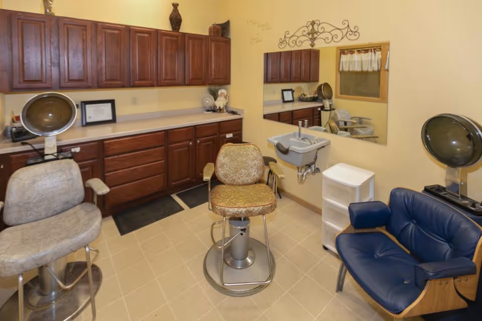 Interior of a salon room with three vintage hair drying chairs, a large mirror on the wall, wooden cabinets, a small sink, and a blue cushioned chair.