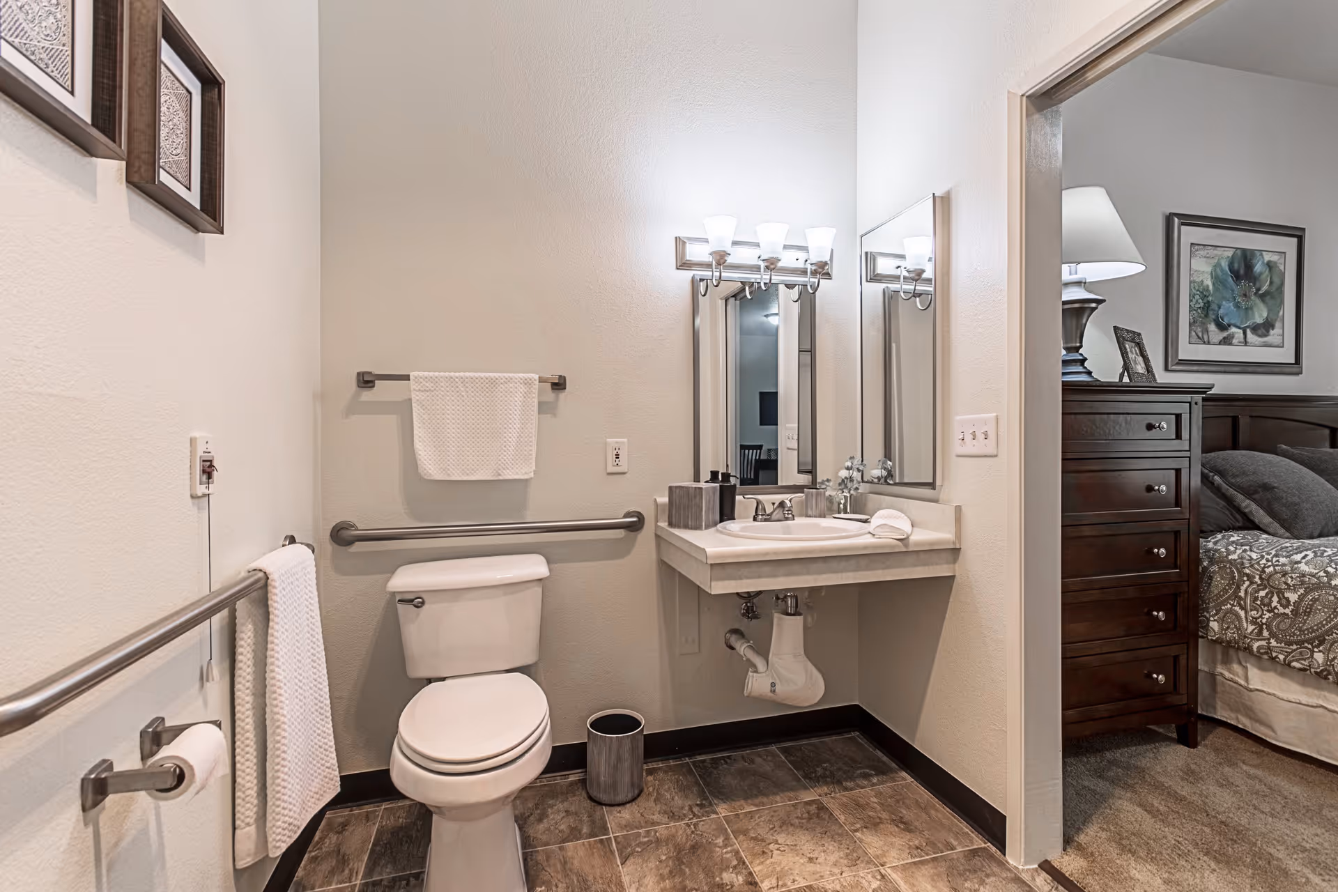 A clean, accessible bathroom with a white toilet, grab bars on the walls, a towel rack with a white towel, a small trash can, and a sink with a large mirror and overhead lighting. The bathroom opens into a bedroom with a wooden dresser, a bed with patterned bedding, a lamp, and framed artwork on the wall.