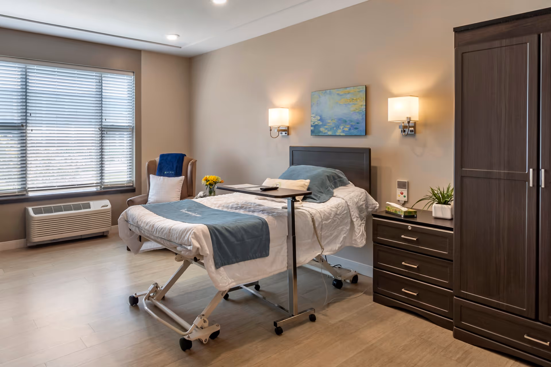 A well-lit senior living bedroom with a hospital-style adjustable bed covered in white and blue bedding. The room features a large window with blinds, a comfortable armchair with a blue throw and white pillow, a bedside table with a plant and tissue box, a dark wood wardrobe, and two wall-mounted lamps. A colorful painting hangs above the bed.