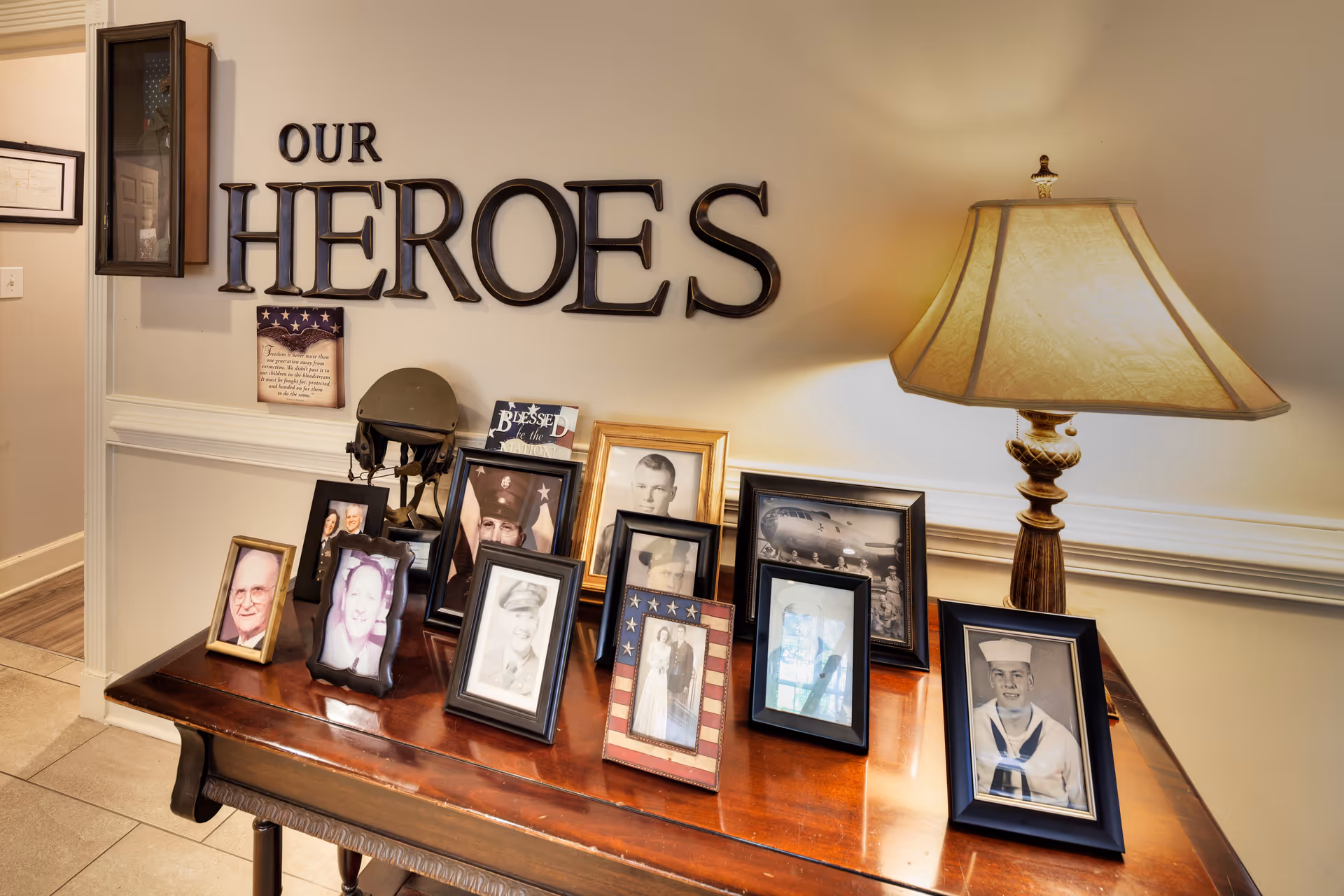 A wooden table displaying framed photographs of veterans and a military helmet, with a lamp on the right side. Above the table on the wall are large letters spelling out 'OUR HEROES'.