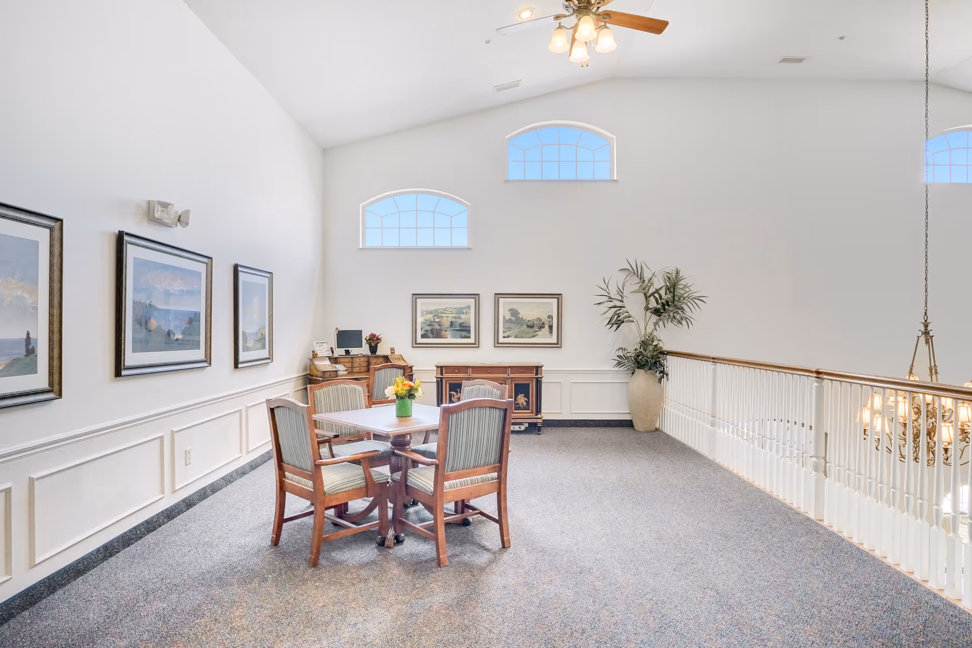 A bright and spacious interior common area with a small square table and four wooden chairs with striped cushions. The room features high ceilings with three arched windows letting in natural light, framed landscape paintings on the walls, a ceiling fan with lights, a large potted plant, and a wooden sideboard with decorative items. The area overlooks a staircase with white railings and a chandelier below.
