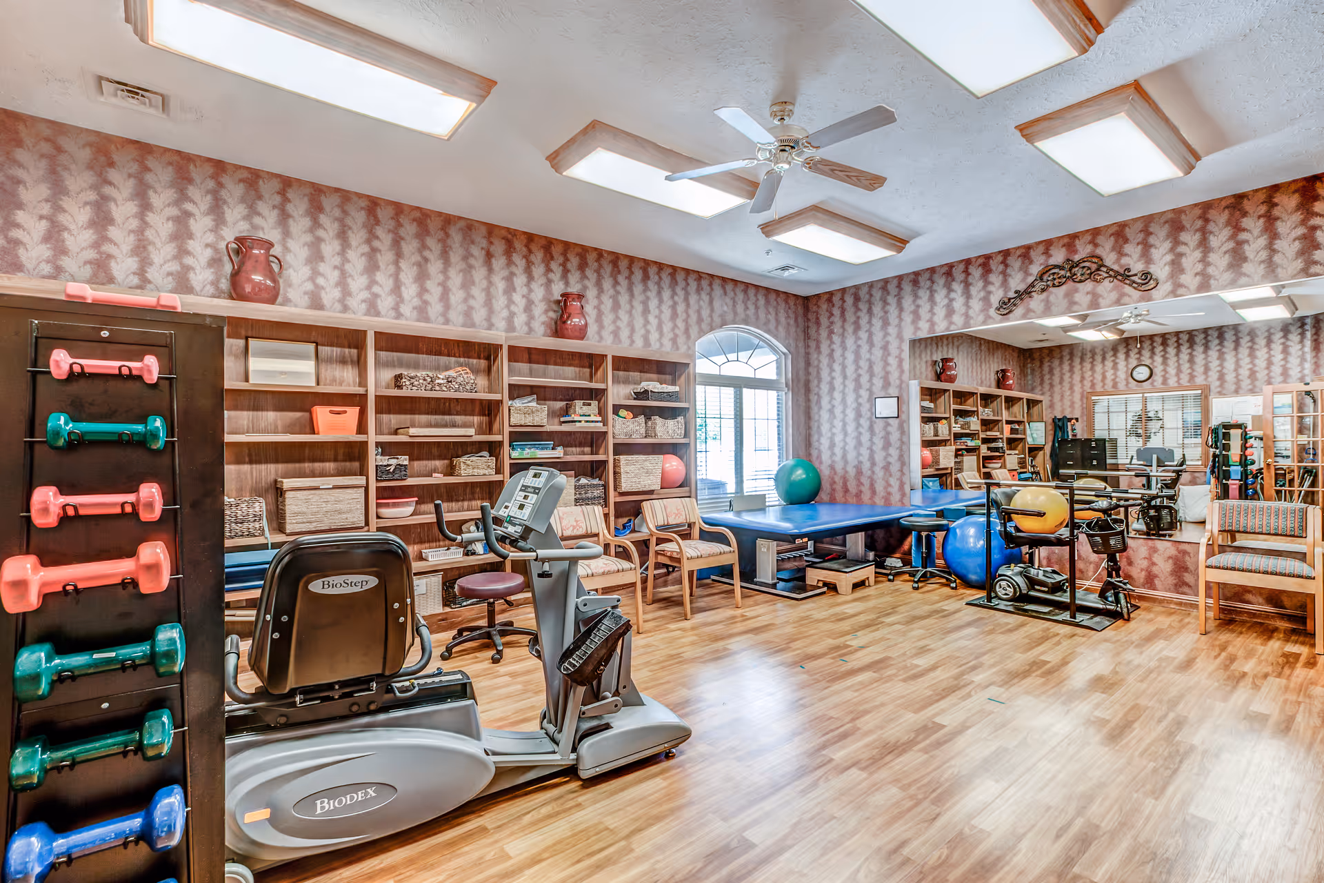 A well-lit exercise and therapy room with wooden flooring, featuring a recumbent exercise bike, colorful dumbbells on a rack, therapy balls, a padded therapy table, chairs, and shelves with baskets and equipment. The room has patterned wallpaper, a large mirror on one wall, and a ceiling fan with multiple fluorescent lights.