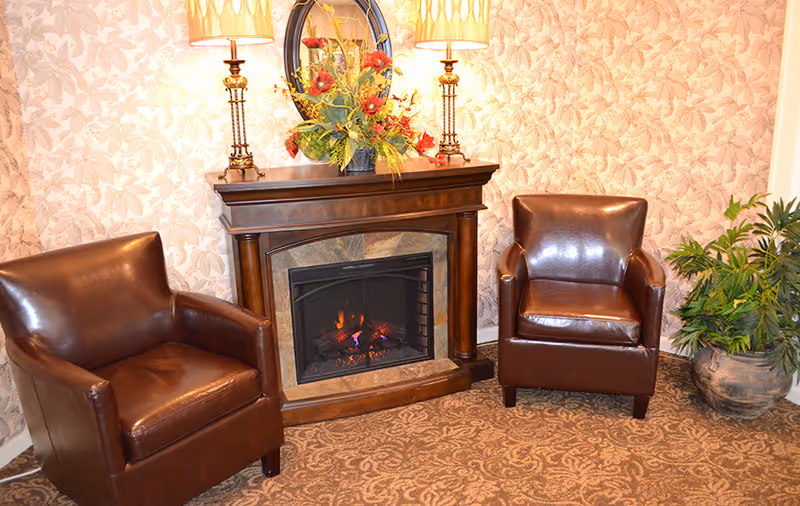 Two brown leather armchairs flanking a lit fireplace mantel decorated with lamps, a mirror, and a floral arrangement in a cozy patterned room.