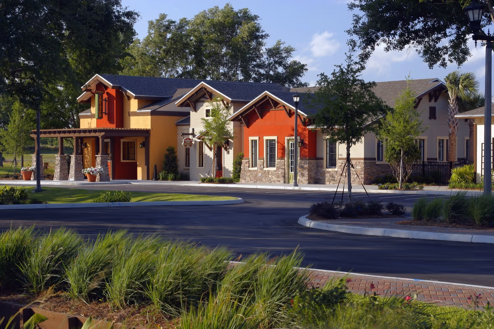 Exterior view of a single-story building with a combination of beige, red, and yellow walls, stone accents, and a dark roof. The building is surrounded by trees, shrubs, and a paved driveway with landscaped greenery in the foreground under a clear sky.