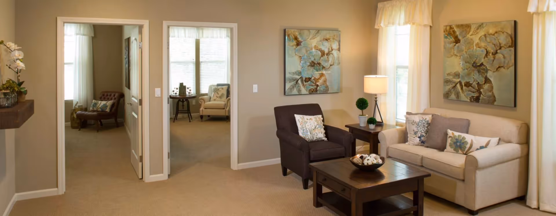A cozy living room with a beige sofa and a dark armchair, both adorned with floral-patterned pillows. A wooden coffee table with a decorative bowl sits in front of the seating area. Two large floral paintings hang on the walls, and a side table with a lamp and small potted plants is next to the armchair. The room has beige walls and carpet, with two doorways leading to other rooms furnished with chairs and windows with sheer curtains.