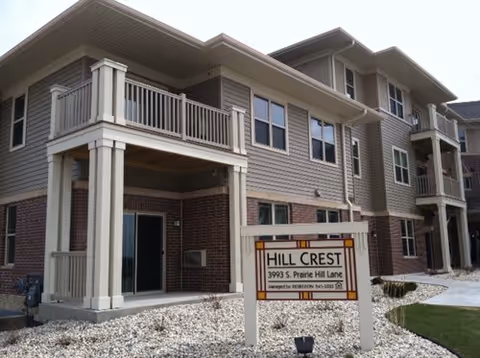 Exterior view of a multi-story residential building with beige siding and brick accents. The building has balconies and multiple windows. In front of the building, there is a sign that reads 'Hill Crest 3993 S. Prairie Hill Lane' with contact information below. The area around the sign is landscaped with white rocks.