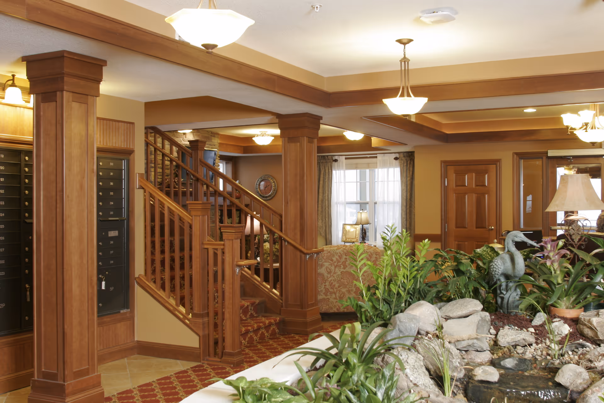Interior view of a senior living facility lobby area featuring wooden columns and staircase with a red patterned carpet. There is a small indoor garden with rocks, plants, and a decorative bird statue in the foreground. The background shows a seating area with upholstered chairs, a window with curtains, and warm lighting fixtures on the ceiling.