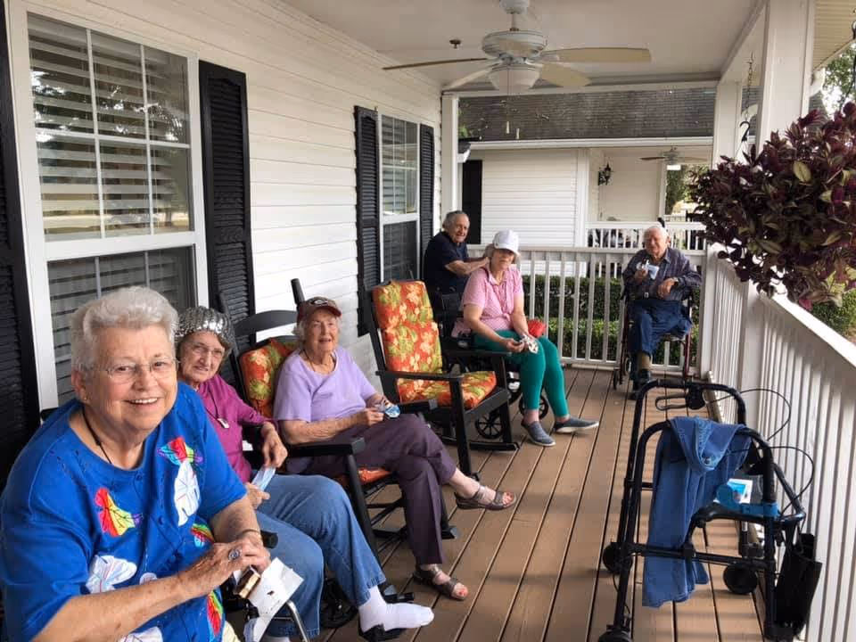A group of six elderly people sitting and relaxing on a covered porch with white railings and black shutters. Some are seated in cushioned rocking chairs, and one person is in a wheelchair. There are walking aids and a hanging plant visible on the porch.