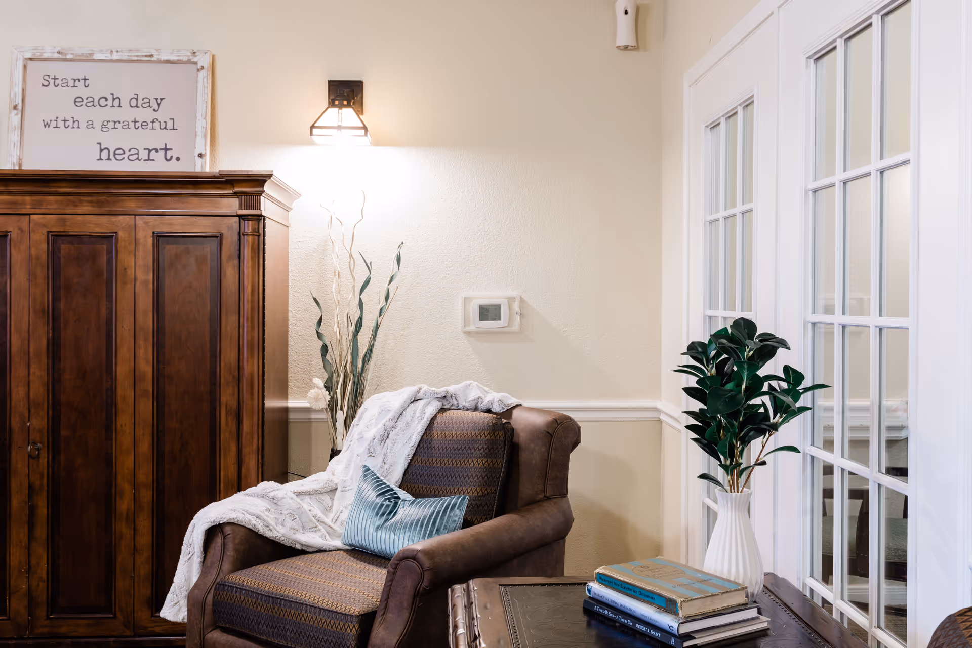 A cozy corner of a living room with a brown upholstered armchair draped with a white blanket and a blue pillow. Next to the chair is a wooden cabinet with a framed sign on top that reads 'Start each day with a grateful heart.' A wall-mounted light fixture illuminates the area. A small table beside the chair holds a white vase with green leafy plants and a stack of books. French doors with glass panes are visible on the right side.