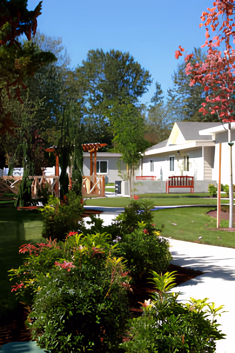 Landscaped outdoor walkway and garden leading to a single-story assisted living building with benches and trees.