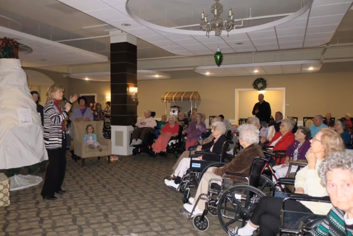 A group of elderly people, many in wheelchairs, seated in a common room listening to a woman speaking. The room has carpeted floors, a chandelier, and holiday decorations including a wreath and a large covered object that appears to be a Christmas tree.