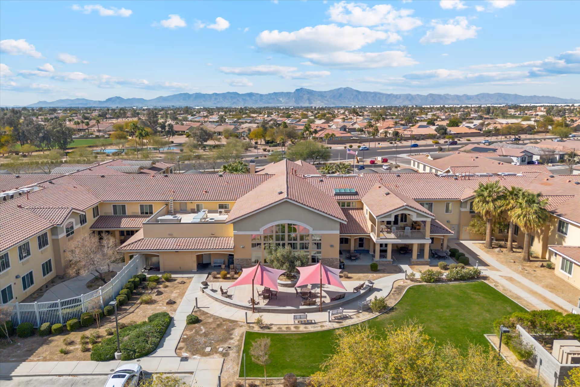 Aerial view of The Groves Assisted Living & Memory Care facility showing a large building with a red-tiled roof, outdoor seating area with pink shade sails, green lawn, and surrounding residential neighborhood with mountains in the background under a partly cloudy sky.