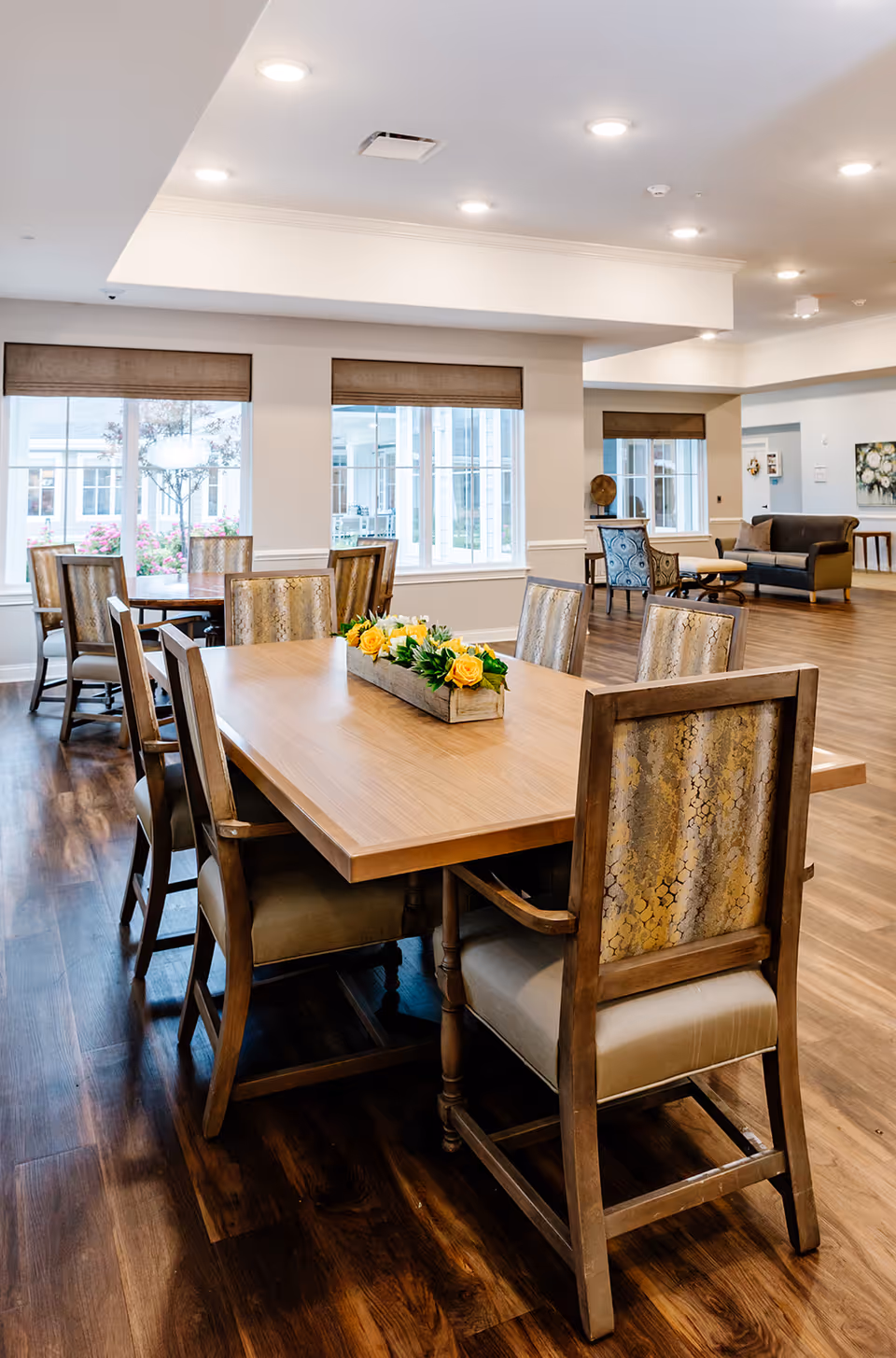 A bright and spacious dining room with a large wooden table surrounded by eight upholstered chairs. The table has a rectangular wooden planter with yellow flowers as a centerpiece. The room features large windows with brown blinds, wooden flooring, and recessed ceiling lights. In the background, there is a seating area with a sofa and armchairs.