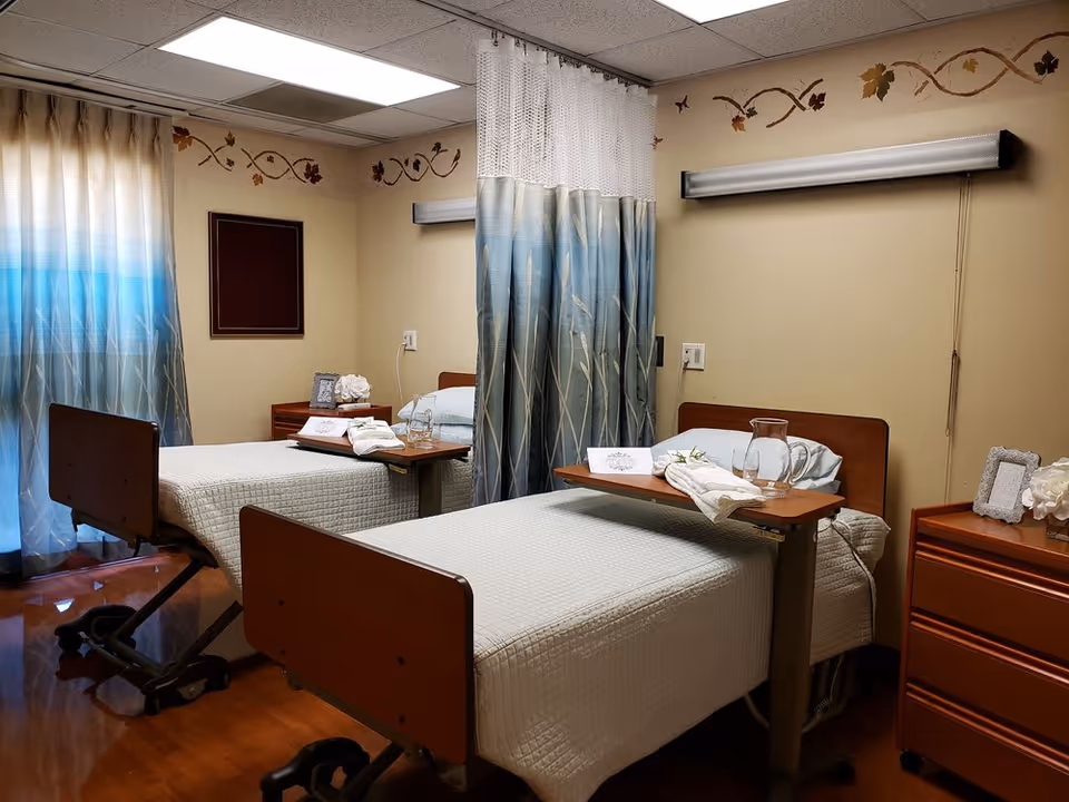 A room with two hospital-style beds separated by a privacy curtain. Each bed has a wooden headboard and footboard, white bedding, and an overbed table with a glass pitcher, glass, and folded towel. The room has beige walls with a decorative leaf border near the ceiling, a wooden nightstand with a framed photo and flowers, and blue gradient curtains covering the window.
