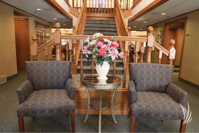 Two patterned armchairs with a small round glass table between them holding a white vase with pink and white flowers. Behind the chairs is a carpeted staircase with wooden railings leading to an upper floor. The setting appears to be a cozy indoor common area with bookshelves visible in the background.