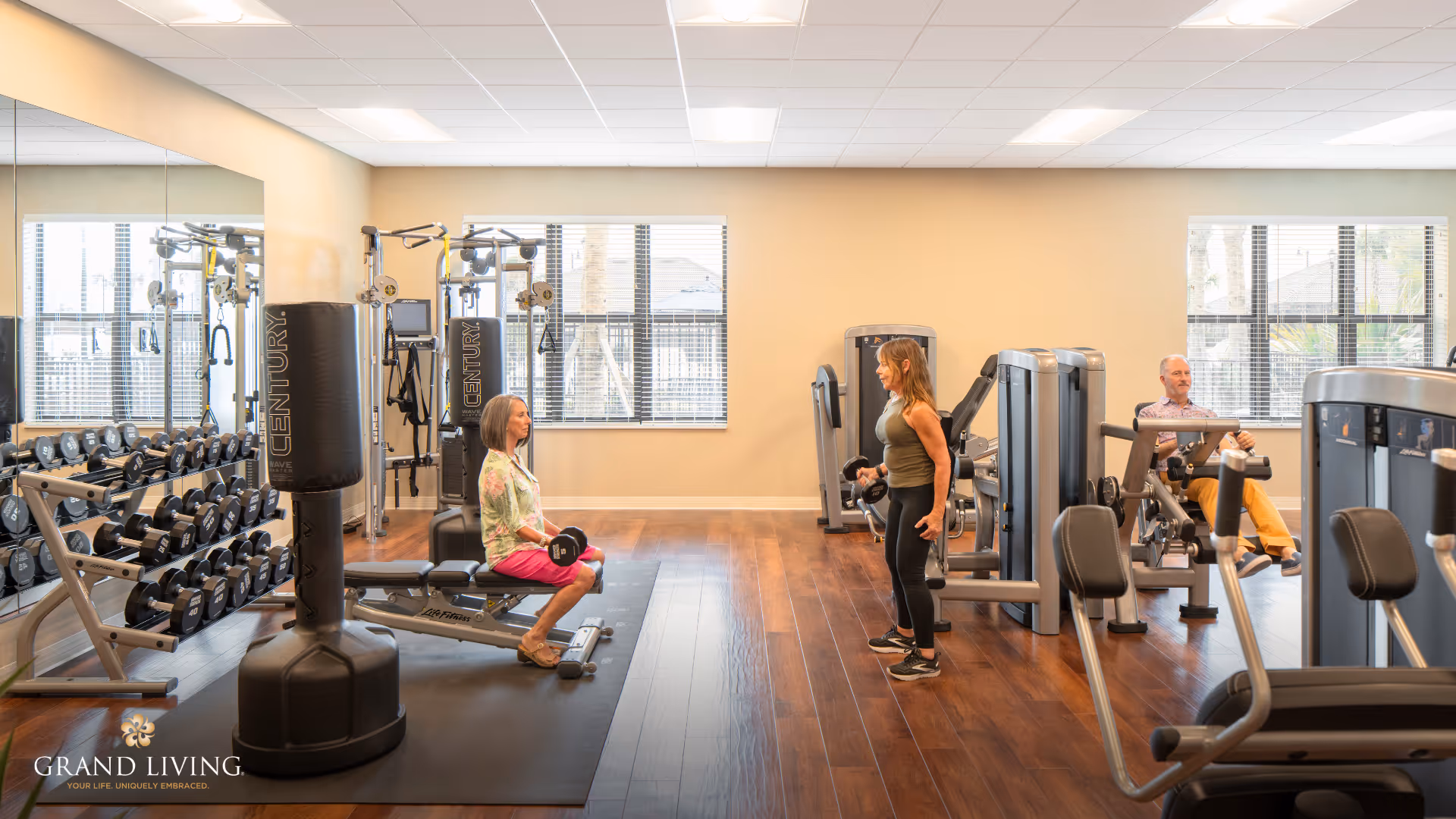 A fitness room with exercise equipment including dumbbells, weight machines, and a punching bag. Three seniors are using the equipment: one woman sitting on a bench holding dumbbells, another woman standing and exercising with dumbbells, and a man seated on a weight machine. The room has large windows, wooden floors, and a mirrored wall.