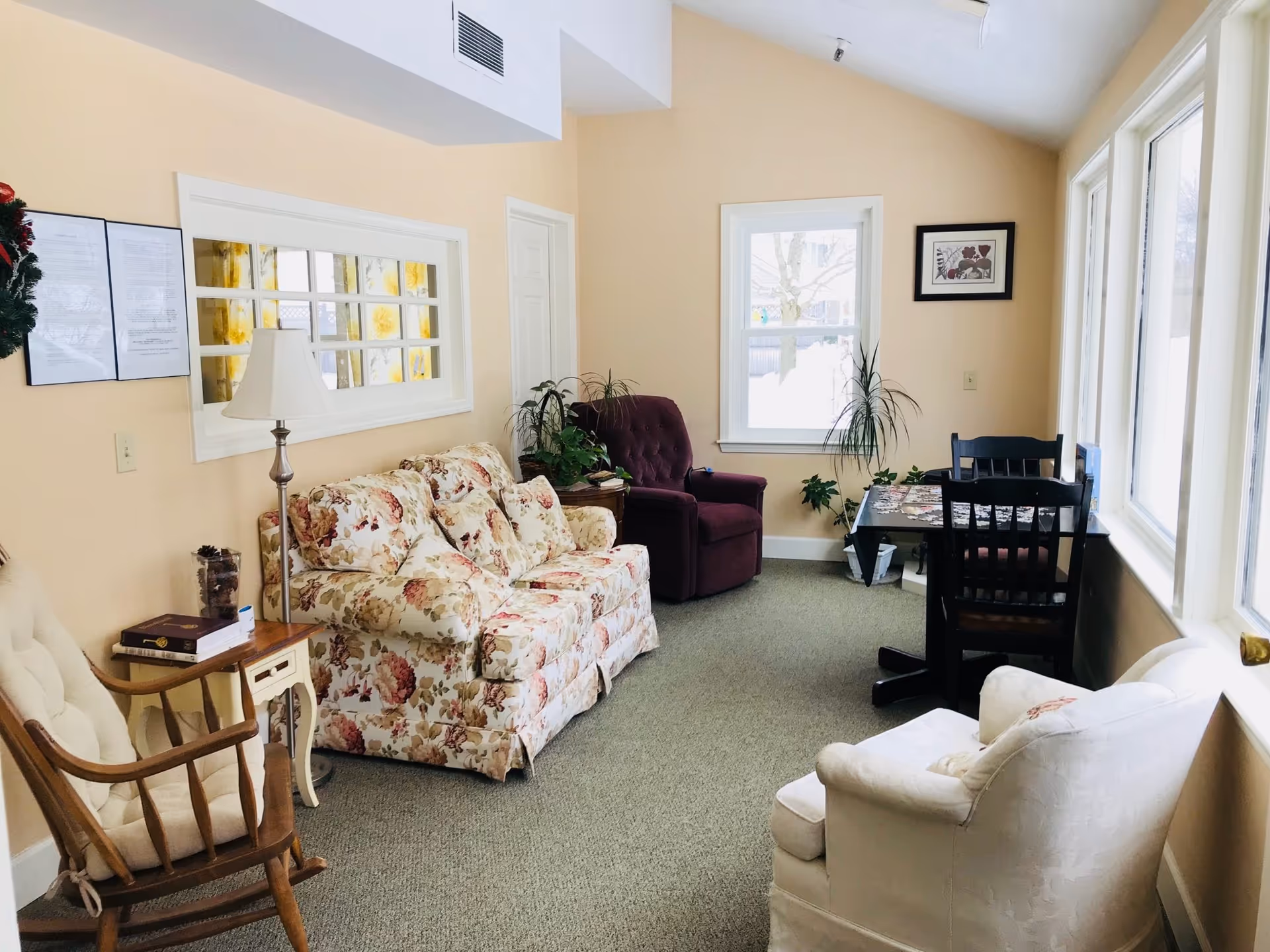 Sunlit communal sitting room with a floral loveseat, several armchairs, a small table and chairs, and large windows.
