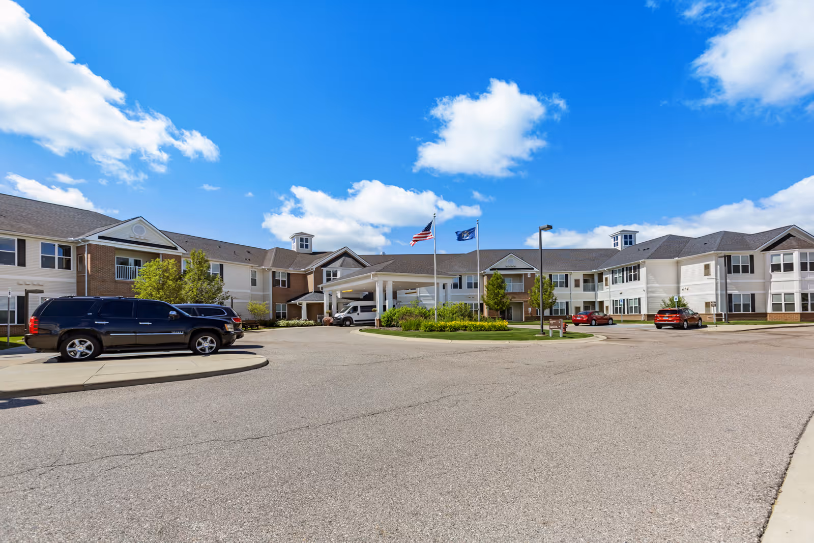 Exterior view of Olivia's Assisted Living facility showing a large two-story building with multiple windows, a covered entrance with two flagpoles flying the American flag and another flag, several parked cars, and a clear blue sky with some clouds.