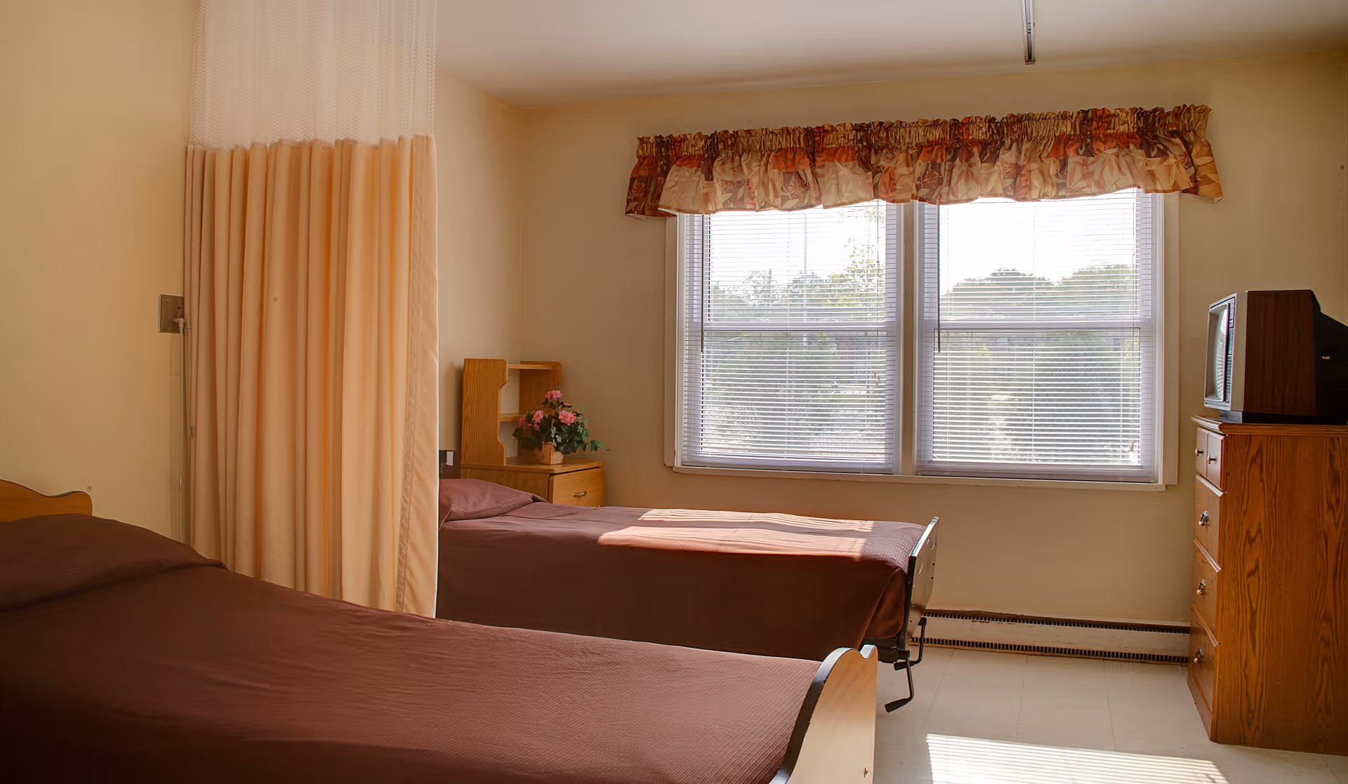 A simple bedroom in a senior living facility with two single beds covered in brown bedspreads, a wooden nightstand with a small flower arrangement, a wooden dresser with an old-style television on top, and a large window with blinds and a floral valance letting in natural light.