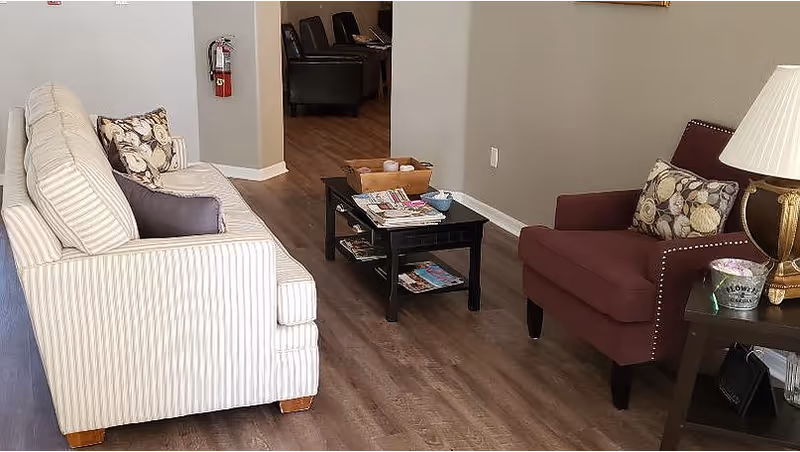 A cozy living room area with a striped beige sofa and a maroon armchair, both adorned with patterned cushions. A black coffee table with magazines and decorative items sits between them on a wooden floor. A side table with a lamp and a small flower pot is next to the armchair. A fire extinguisher is mounted on the wall in the background.