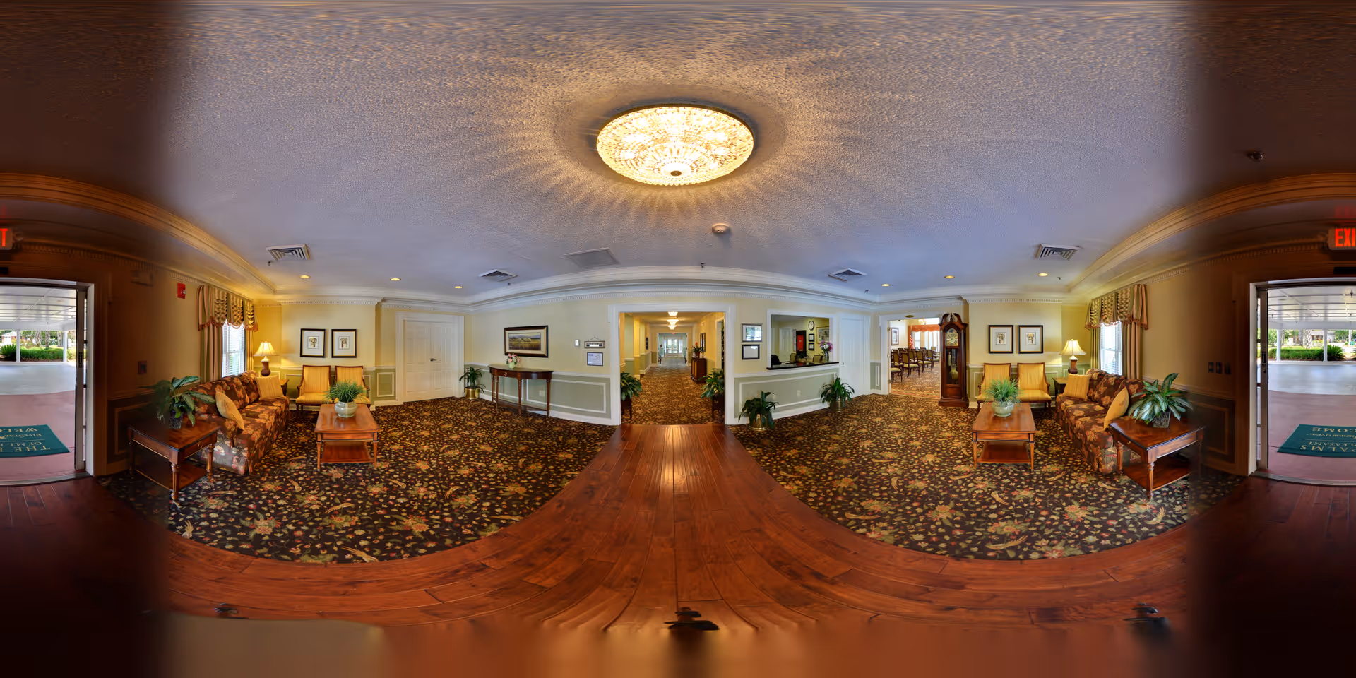 A spacious and warmly decorated senior living facility lobby with patterned carpet, wooden flooring, and comfortable seating areas including sofas and armchairs. There are plants on tables and framed artwork on the walls. The lobby has two open doors leading outside, and a hallway leading further into the building. A large ceiling light fixture illuminates the area.