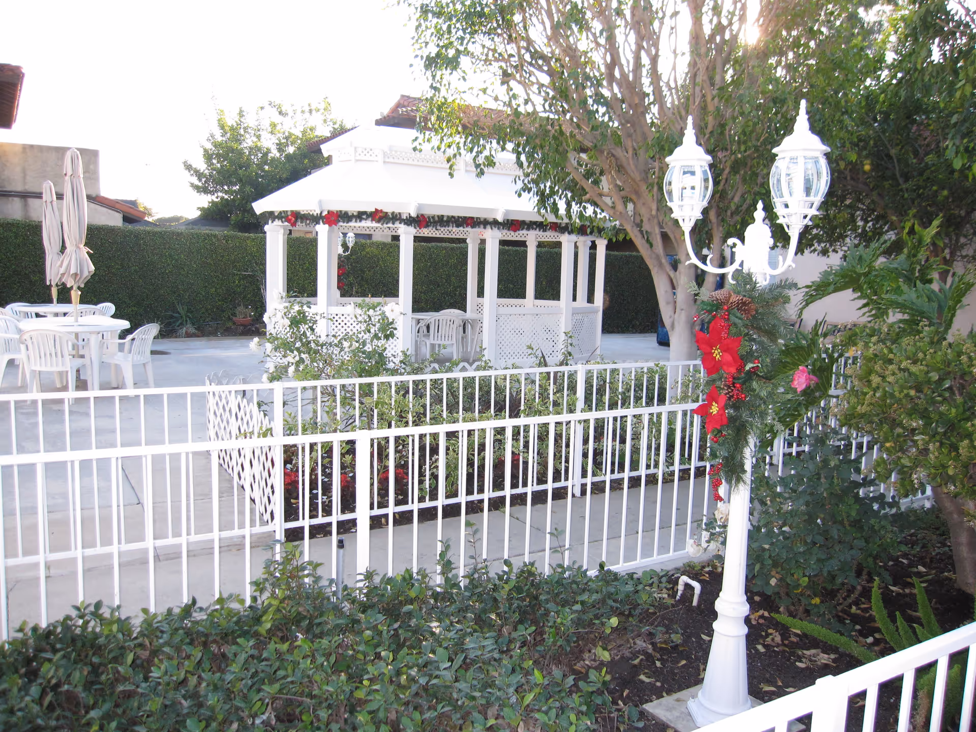 Outdoor garden area with a white gazebo decorated with red flowers, white plastic chairs and tables with closed umbrellas, a white metal fence, and a white lamp post adorned with festive red poinsettia decorations and greenery. Surrounding the area are trees and bushes.