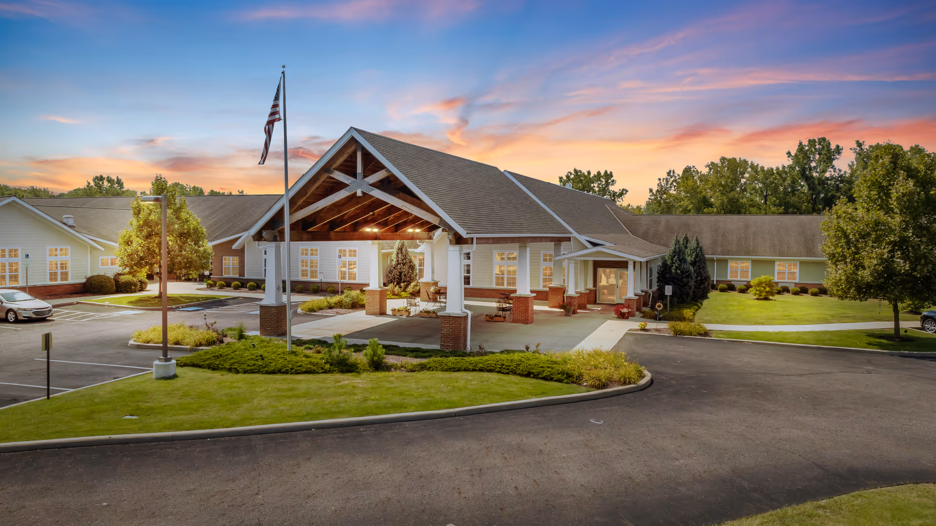 Front entrance of a single-story senior care building with a covered porte-cochere, flagpole, parking lot, and landscaped grounds at sunset.