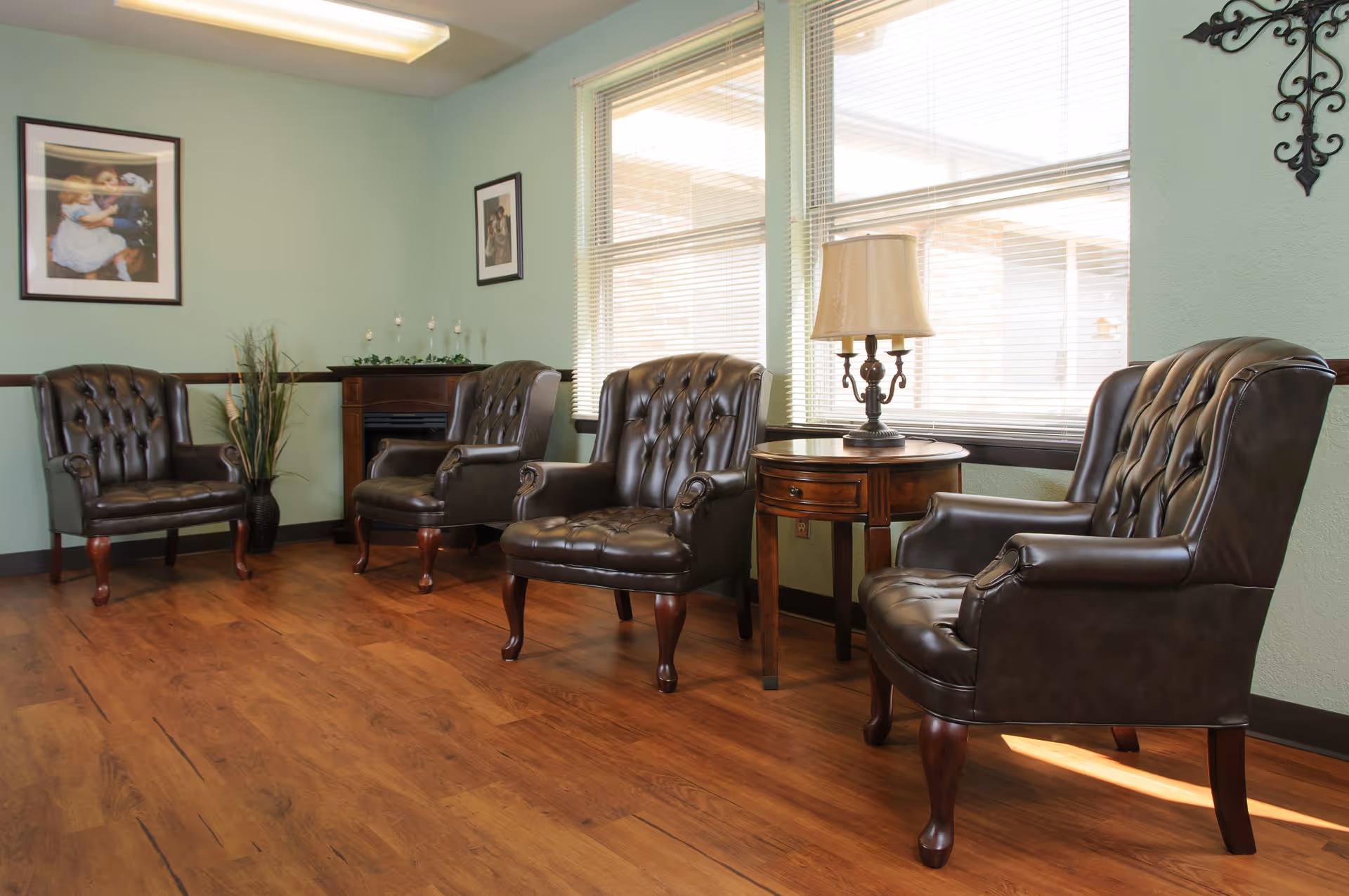 A cozy sitting area with four dark brown leather armchairs arranged around a wooden side table with a lamp. The room has light green walls, two framed pictures, a small decorative fireplace, and large windows with blinds letting in natural light. The floor is wooden.