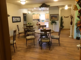 Interior view of a senior living facility dining area with multiple round tables and chairs arranged on a wooden floor. The room is well-lit with ceiling lights and decorated with plants and framed pictures on the walls. In the background, there is a kitchen area with light-colored cabinets and a countertop.