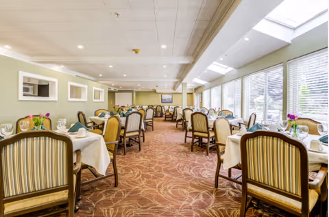 A bright dining room with multiple tables covered with white tablecloths, each set with plates, glasses, napkins, and small flower arrangements. The room has large windows on one side letting in natural light, patterned carpet flooring, and comfortable upholstered chairs around the tables.