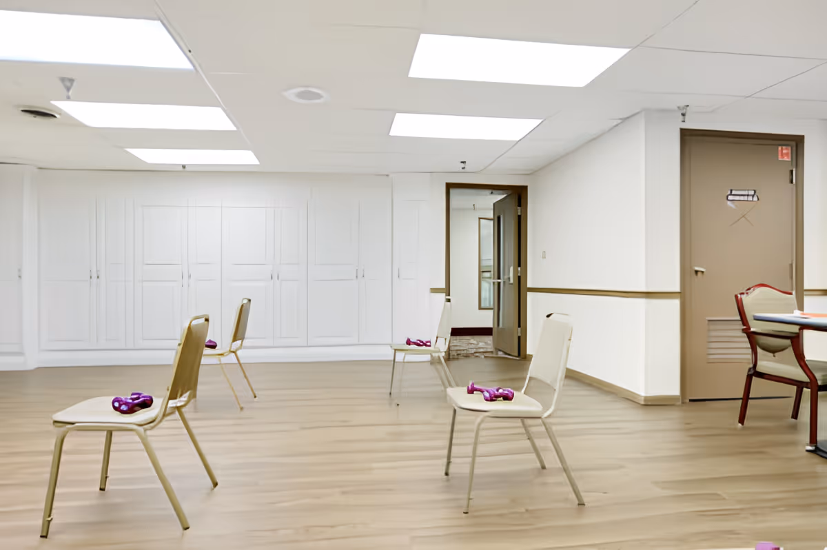 A spacious room with wooden flooring and white walls, featuring four beige chairs spaced apart, each with a pair of small purple dumbbells placed on the seat. The ceiling has recessed lighting panels, and there is a closed brown door and an open door leading to another room. A table with chairs is partially visible on the right side.