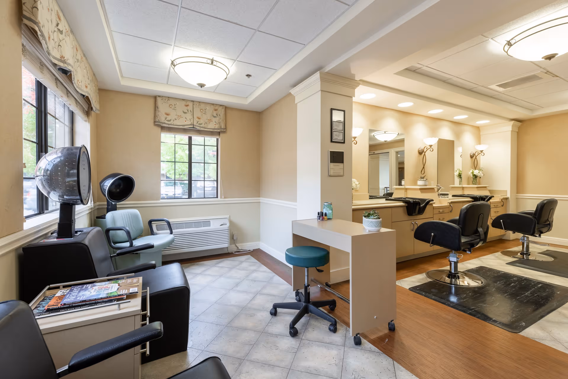 Interior view of a senior living facility hair salon area with two black salon chairs in front of mirrors and sinks, a small workstation with a green stool, and a waiting area with chairs and hair drying machines near windows.
