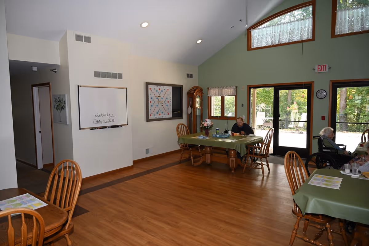 Interior of a senior living facility dining area with wooden floors and green walls. Several wooden tables covered with green tablecloths are arranged around the room, with wooden chairs placed around them. Two elderly individuals are seated at separate tables, one in a wheelchair. A whiteboard on the wall displays the date Saturday, October 2nd, 2021. Large windows and glass doors let in natural light and provide a view of an outdoor patio with chairs and greenery.