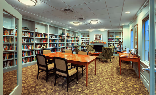 A cozy library room with bookshelves filled with books lining the walls. In the center, there is a wooden table with six chairs around it. At the far end of the room, there are two upholstered armchairs facing a fireplace with a framed picture above it. The room has patterned carpet flooring, soft ceiling lights, and windows on the right side letting in natural light.