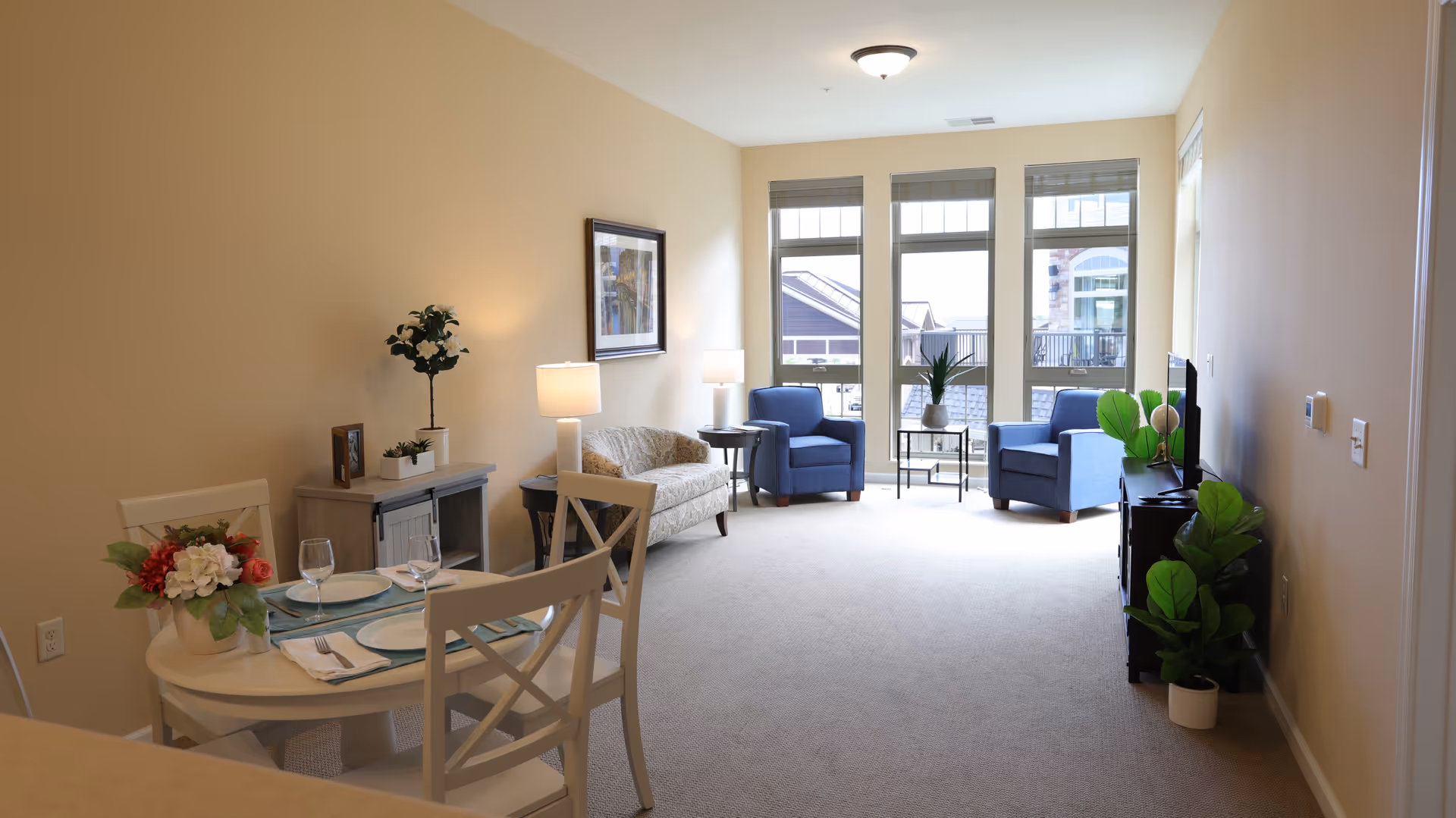Bright open living and dining area with a small round dining table, sofa, two blue armchairs and large windows letting in natural light.
