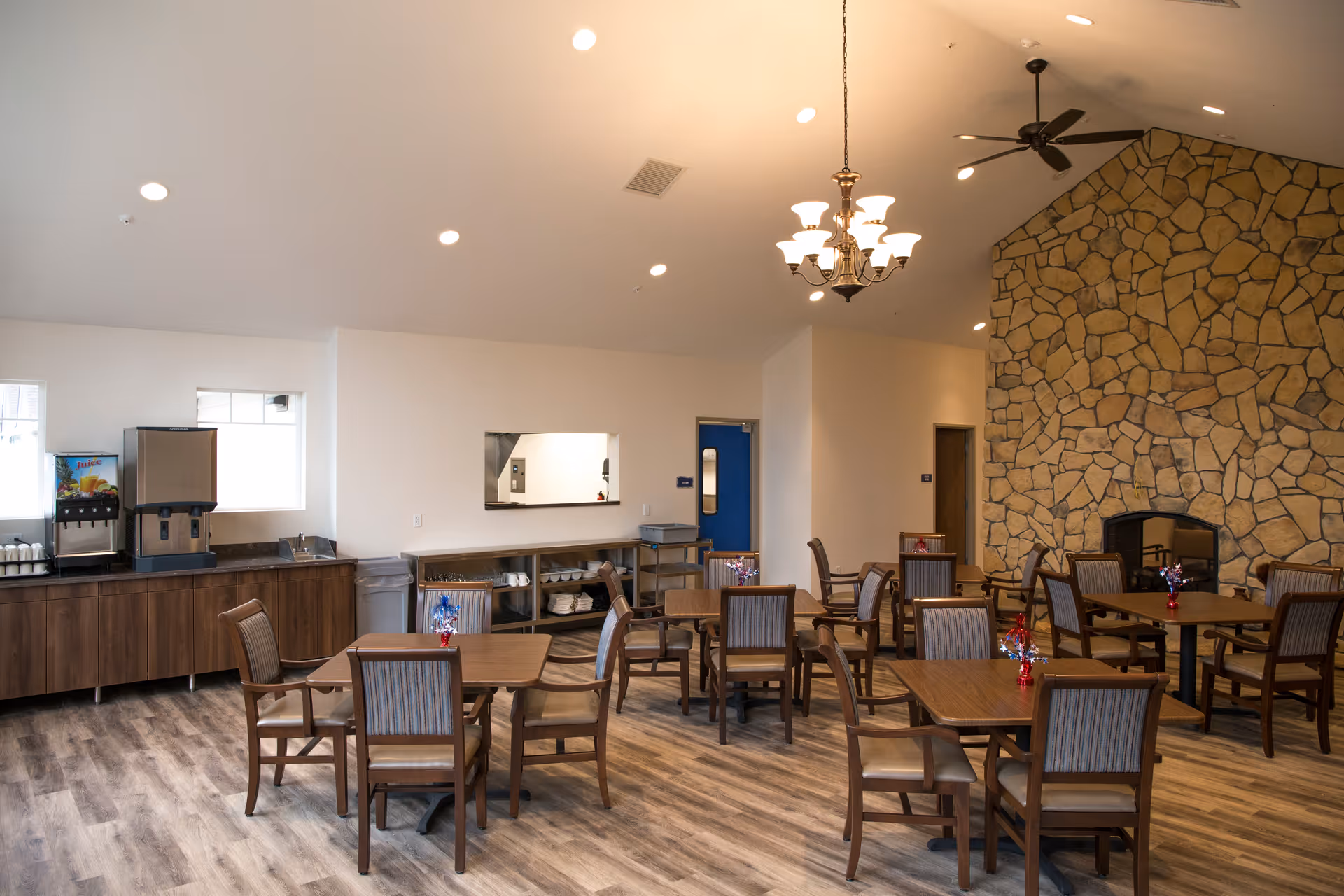 Spacious dining room with multiple wooden tables and chairs, a stone fireplace wall, and a beverage station along one side.