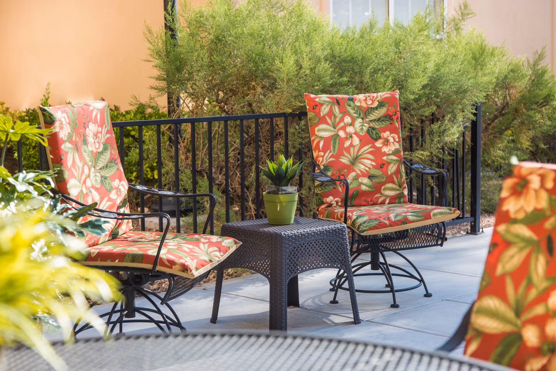 Outdoor patio area with two metal chairs featuring red floral cushions and a small black wicker table with a green potted plant on top. Green bushes and a black metal fence are visible in the background.