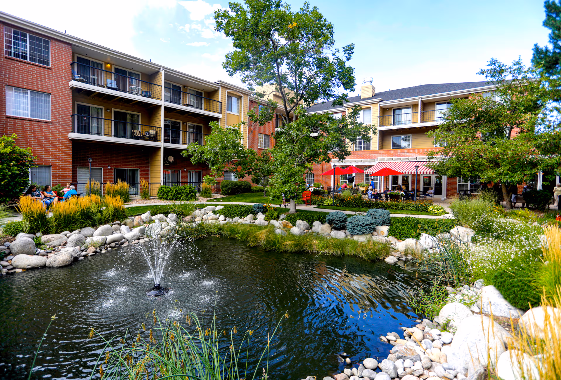 Courtyard view of a multi-story retirement building with a landscaped pond and fountain, patio seating and red umbrellas.