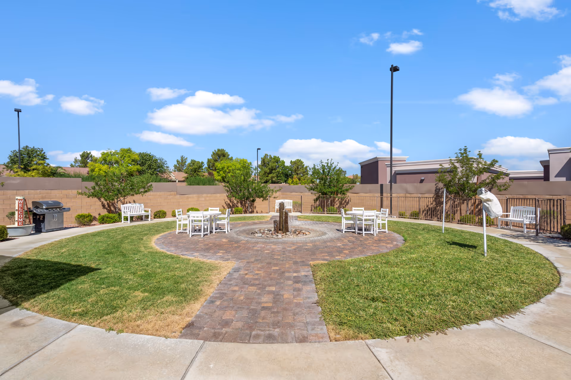 Sunny circular courtyard with a brick walkway, central water feature, white tables and benches, and a barbecue grill.