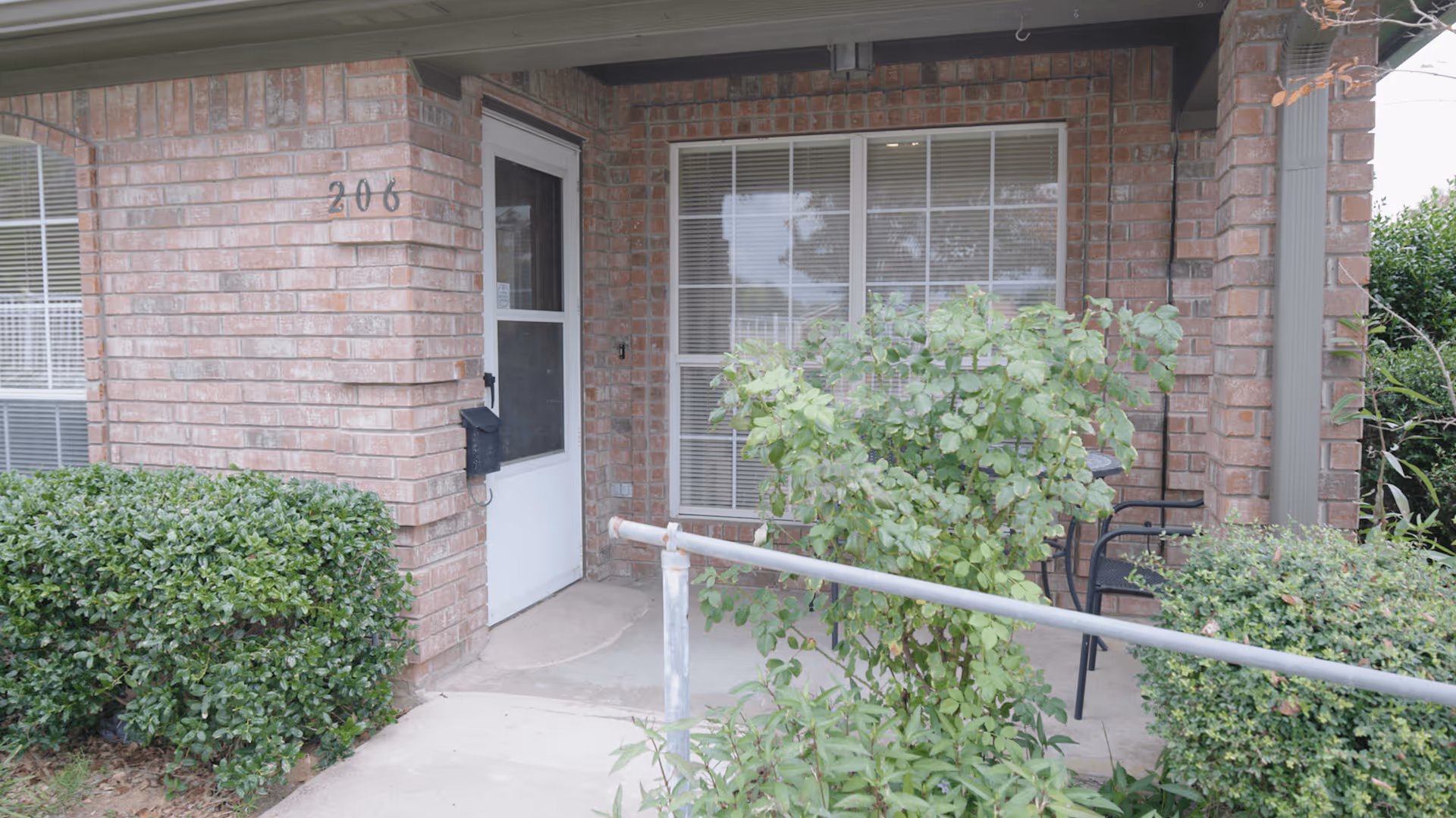 Front porch of a brick building with the number 206 displayed on the wall. The porch has a white door with a screen, a large window with blinds, green bushes, a metal handrail, and a black chair partially visible behind the bushes.