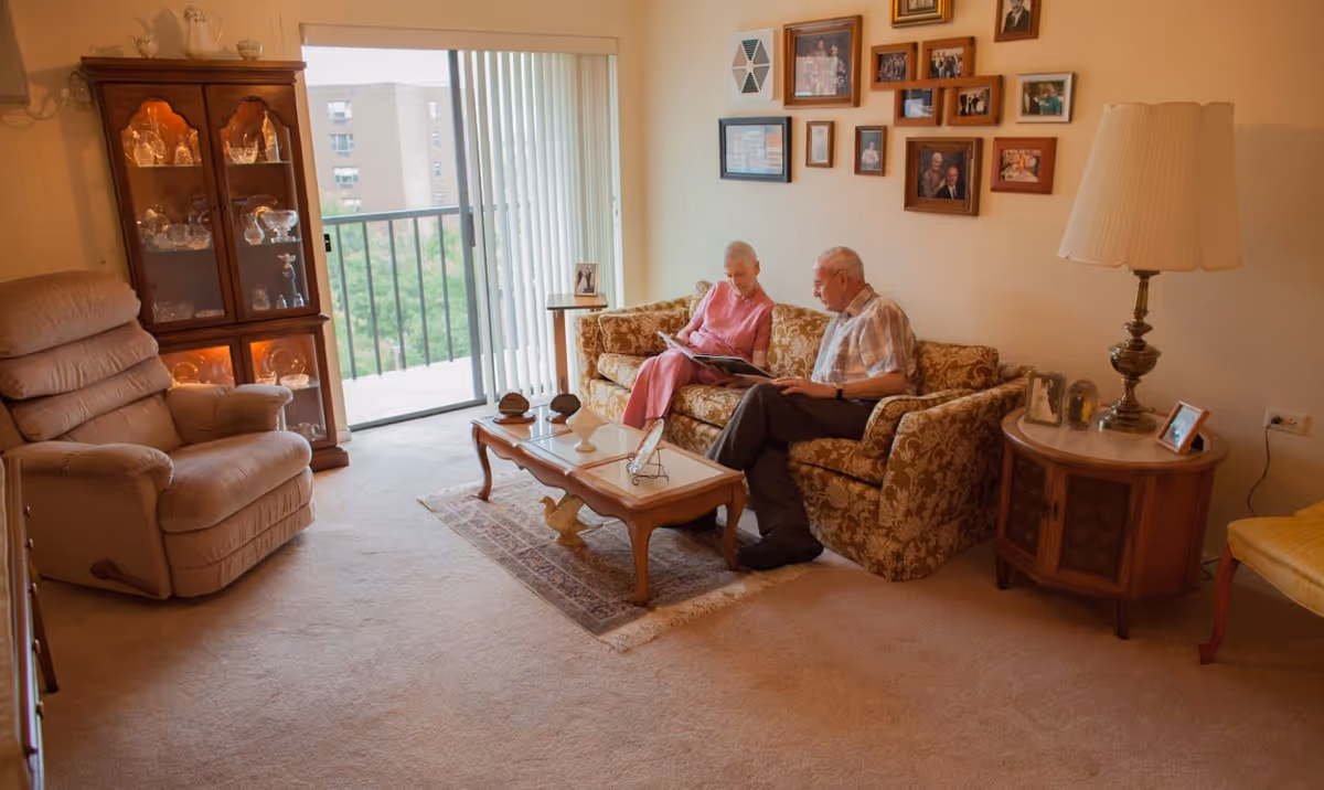An elderly couple sitting on a patterned sofa in a cozy living room, looking at a photo album. The room features a glass-front wooden cabinet with decorative glassware, a beige recliner chair, a wooden coffee table with decorative items, a side table with a lamp and framed photos, and a wall adorned with multiple framed pictures. A sliding glass door with vertical blinds leads to a balcony with a view of other buildings and greenery outside.