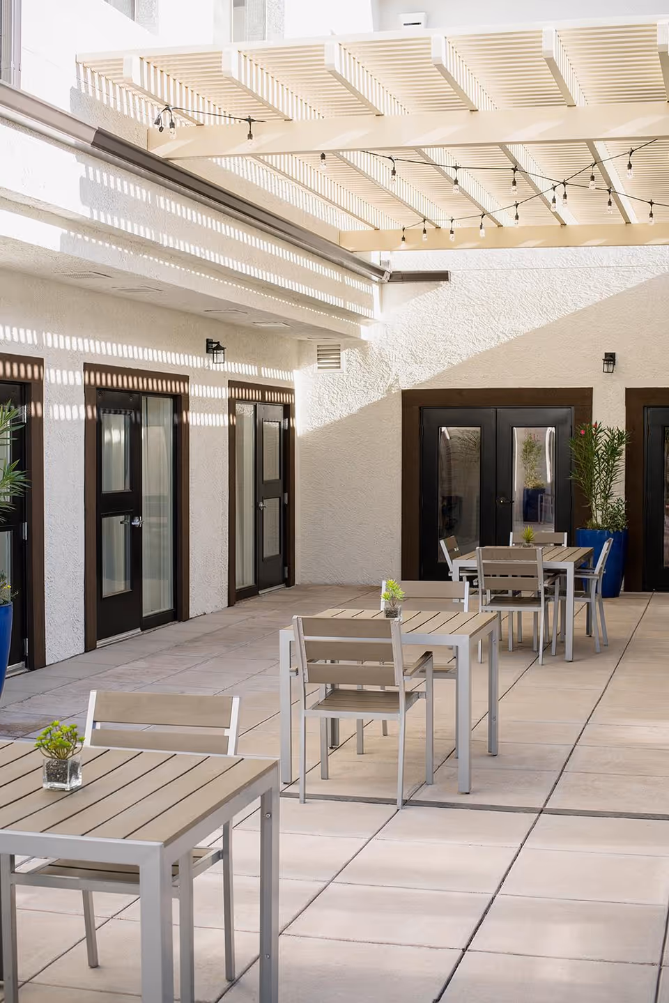 Sunlit courtyard with small patio tables and chairs under a slatted pergola beside doors and potted plants.