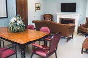 A senior living common room featuring a wooden table with a floral centerpiece and red chairs, plus a seating area with a sofa and armchairs facing a fireplace with a TV above.