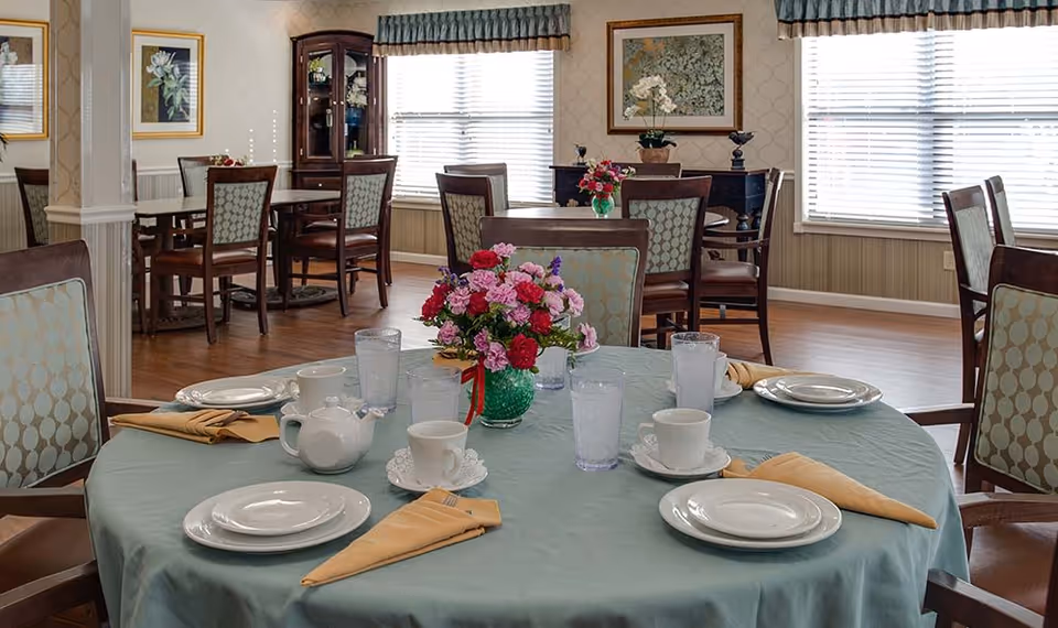 A dining room in a senior living facility with a round table set for four people. The table has a light blue tablecloth, white plates, cups, glasses of water, a teapot, and beige napkins folded neatly. A green vase with a colorful bouquet of flowers is in the center. In the background, there are more tables and chairs, large windows with blinds, framed floral artwork on the walls, and wooden flooring.
