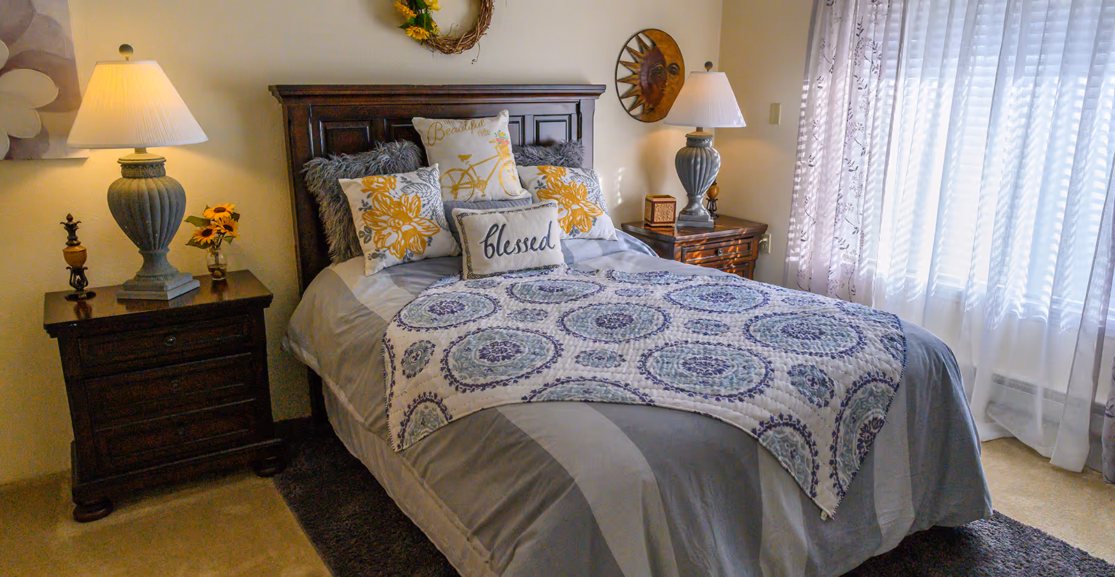 A cozy bedroom with a neatly made bed featuring multiple decorative pillows, including one with the word 'blessed' and another with a bicycle design. The bed has a patterned quilt and gray bedding. On either side of the bed are wooden nightstands with matching lamps, a small vase with sunflowers on the left nightstand, and a decorative sun wall hanging above the right nightstand. Sheer curtains allow natural light to fill the room.
